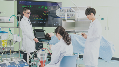 ベッドに横たわった人体模型を用いて装置の使い方を学ぶ男子生徒と女子生徒